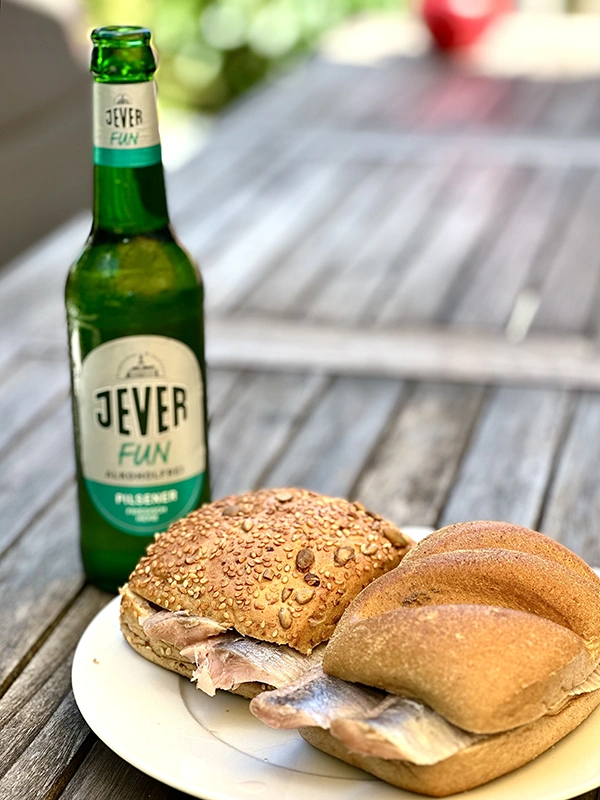 Sonnenuntergang am Meer mit zwei Jever Pilsener Flaschen im Vordergrund, umgeben von blühenden Pflanzen und felsiger Küste.