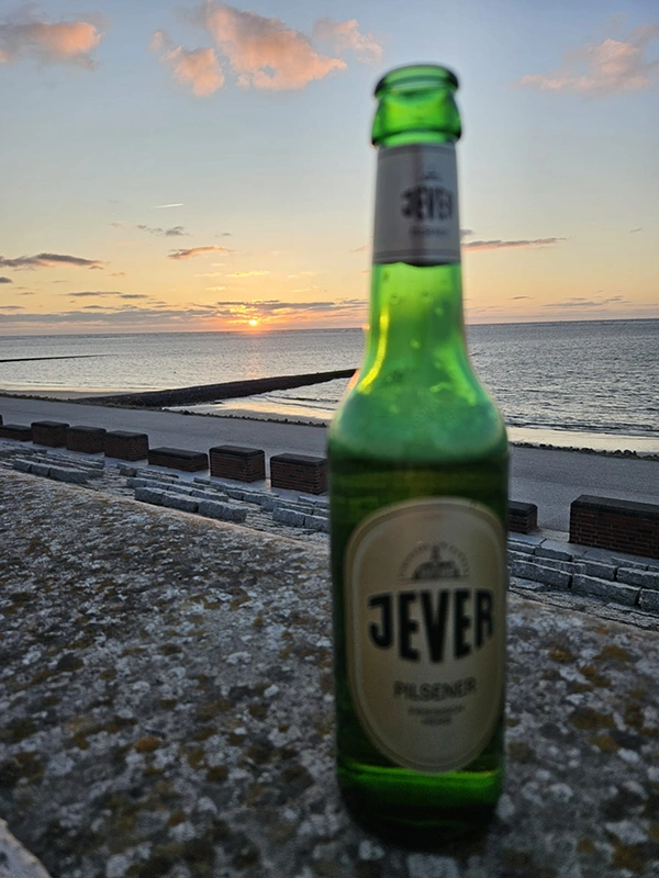 Jever Bierflasche auf einer Mauer, dahinter ein malerischer Sonnenuntergang über dem Meer, Wolken ziehen am Himmel vorbei. 