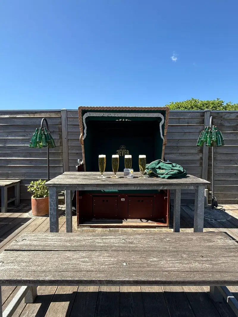 Tisch mit vier Jever Biergläser und Jever-Handtuch vor einem Jever-Strandkorb neben zwei dekorativen Jever-Flaschenlampen.