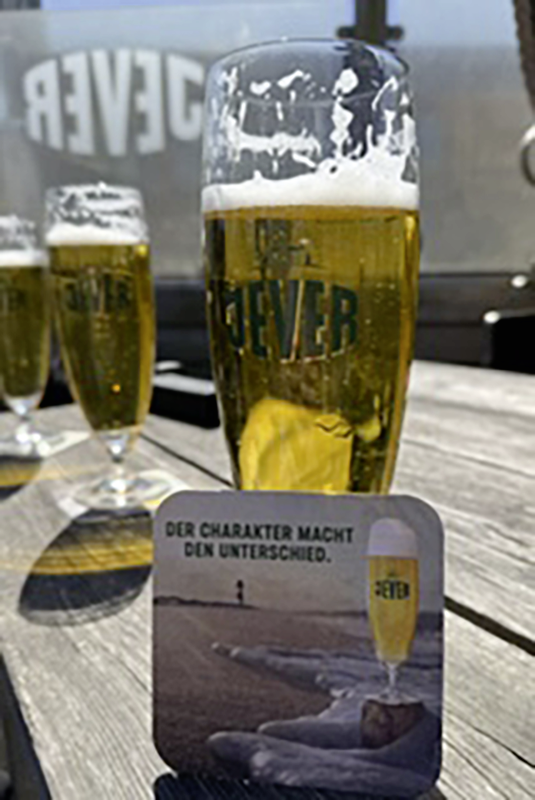Drei Biergläser mit Jever-Logo auf einem Tisch in der Sonne. Im Vordergrund ein Bierdeckel mit Strandmotiv und Werbeslogan.