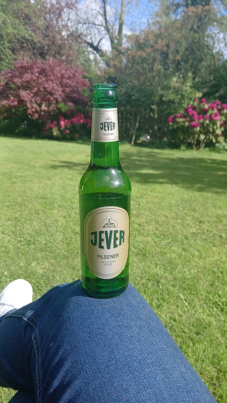 Jever Pilsener Flasche auf einem Knie im Garten. Sonnenschein, grüne Wiese, bunte Blumen und Bäume im Hintergrund.
