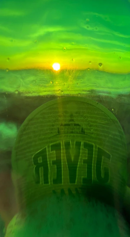Blick durch eine grüne Flasche auf einen Sonnenuntergang. Die Flasche zeigt ein Etikett mit dem Schriftzug 