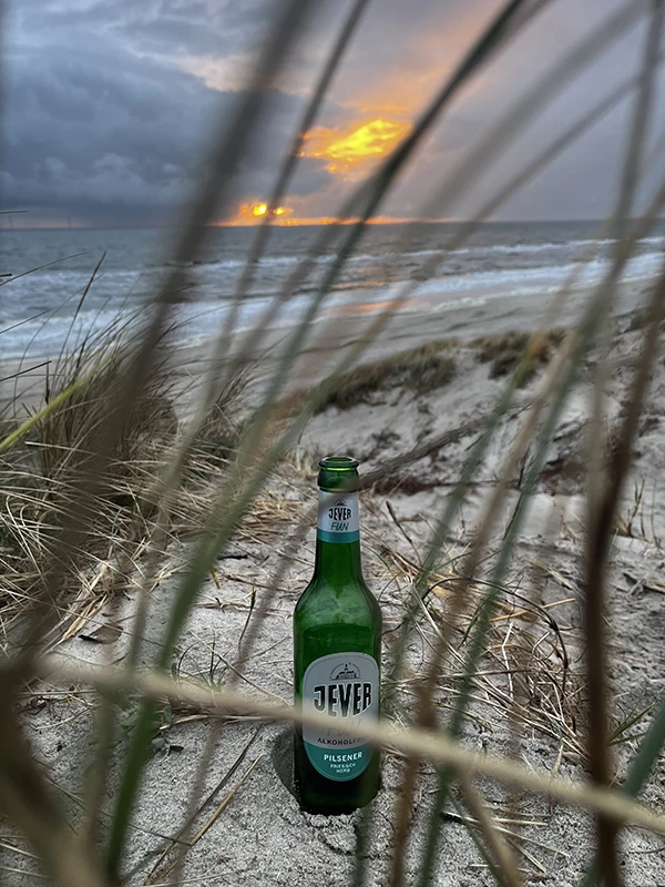 Zwischen Grashalmen am Strand steht eine Flasche Jever Fun. Der Himmel zeigt einen farbenfrohen Sonnenuntergang über dem Meer. 