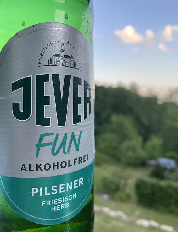 Jever Fun alkoholfreies Pilsener, friesisch herb, vor einer verschwommenen Landschaft mit Bäumen und blauem Himmel im Hintergrund.