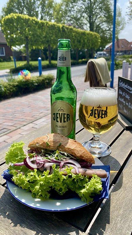 Flasche und Glas Jever Pilsener auf einem Tisch im Freien, daneben ein belegtes Brötchen mit frischem Salat.