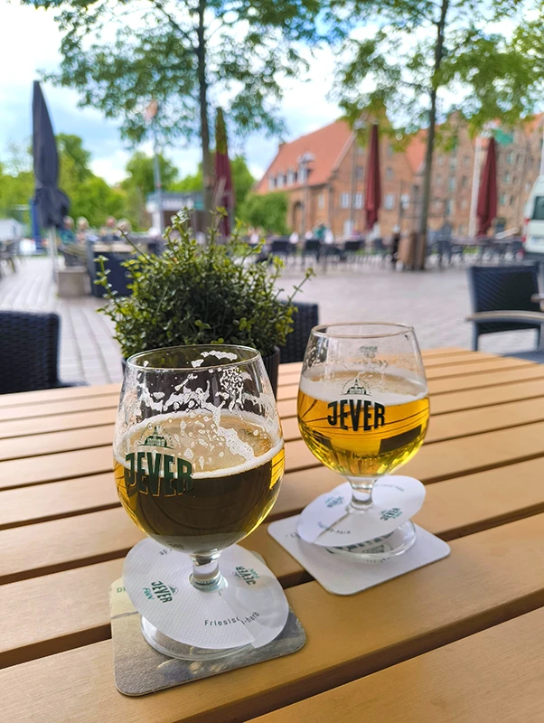 Zwei halbvolle Biergläser auf einem Tisch im Freien, mit einem belebten Platz und historischen Gebäuden im Hintergrund.