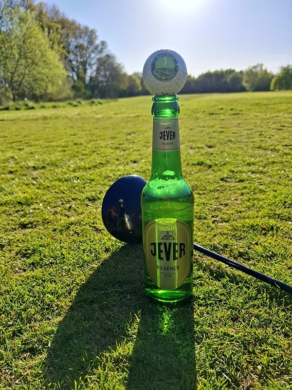 Eine Jever Pilsener Flasche steht auf einem Golfplatz, mit einem Golfball oben drauf und einem Golfschläger daneben.