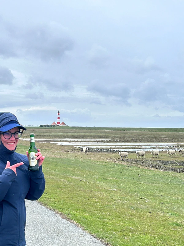 Person in Regenkleidung hält eine Jever Flasche. Im Hintergrund Schafe auf einer Wiese und ein Leuchtturm.