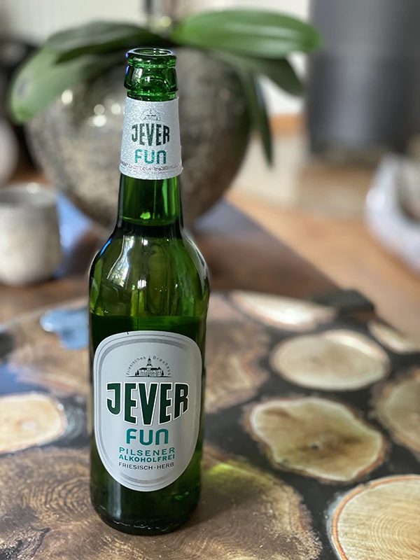 Grüne Jever Fun Pilsener Flasche auf einem Tisch mit Holzstruktur. Im Hintergrund ist eine unscharfe Pflanze zu sehen.