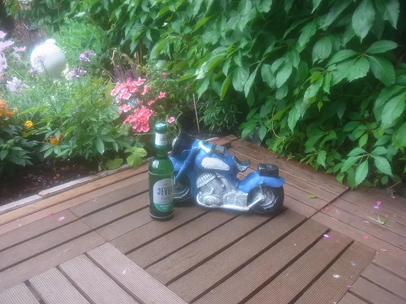 Eine Flasche Jever Fun steht neben einer blauen Motorradfigur auf einer Holzterrasse, umgeben von grünem Laub und Blumen.