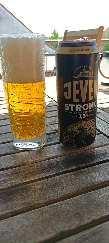 Glas mit hellem Bier neben einer Dose Jever Strong auf einem Holztisch.