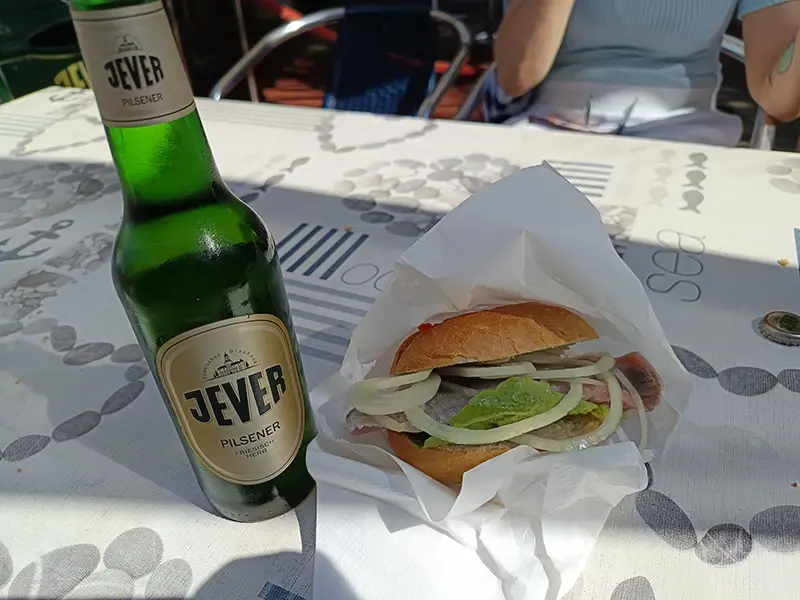 Ein Fischbrötchen mit Zwiebeln und Salat liegt neben einer Flasche Jever Pilsener auf einem Tisch mit maritimem Muster.