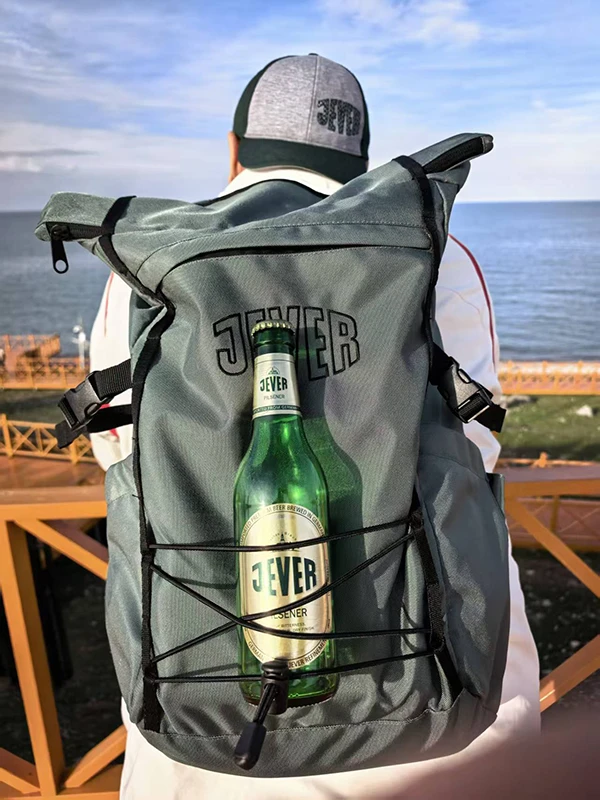 Person mit grauem Rucksack mit Jever-Logo und Jever-Bierflasche steht an einem Geländer, mit Blick auf das Meer.