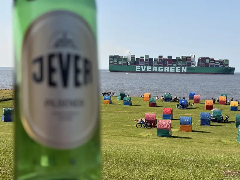 Eine Jever-Bierflasche im Vordergrund, dahinter bunte Strandkörbe auf einer Wiese und ein großes Containerschiff im Meer.