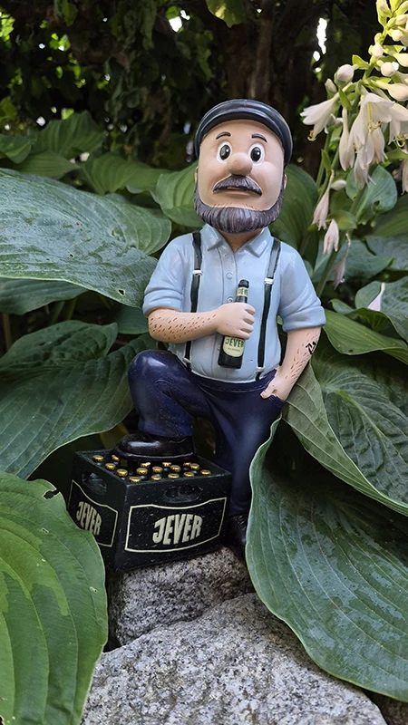 Eine Figur eines Mannes mit Bart und Mütze sitzt auf einem Jever-Bierkasten, umgeben von großen grünen Blättern.