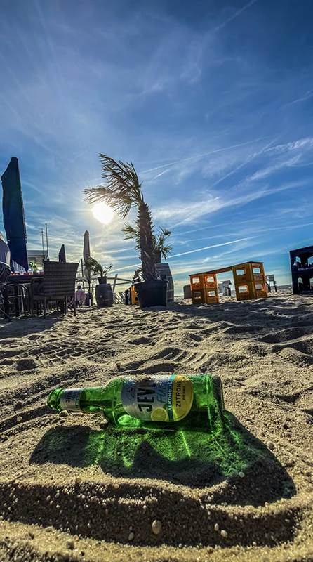 Eine leere Jever-Flasche liegt im Sand eines sonnigen Strandes, umgeben von Palmen und blauen Himmel mit vereinzelten Wolken.