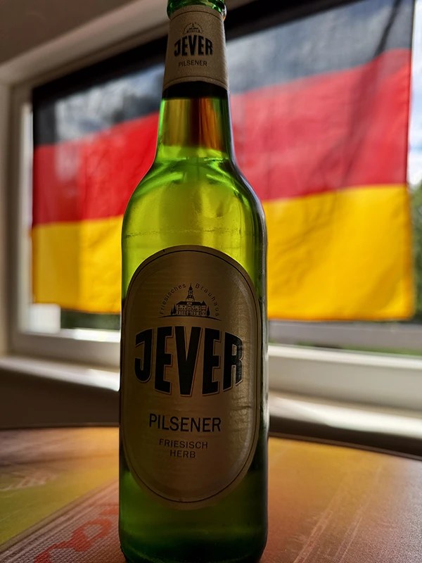 Auf einem Tisch steht eine Jever Bierflasche. Im Hintergrund weht eine deutsche Flagge vor einem Fenster.