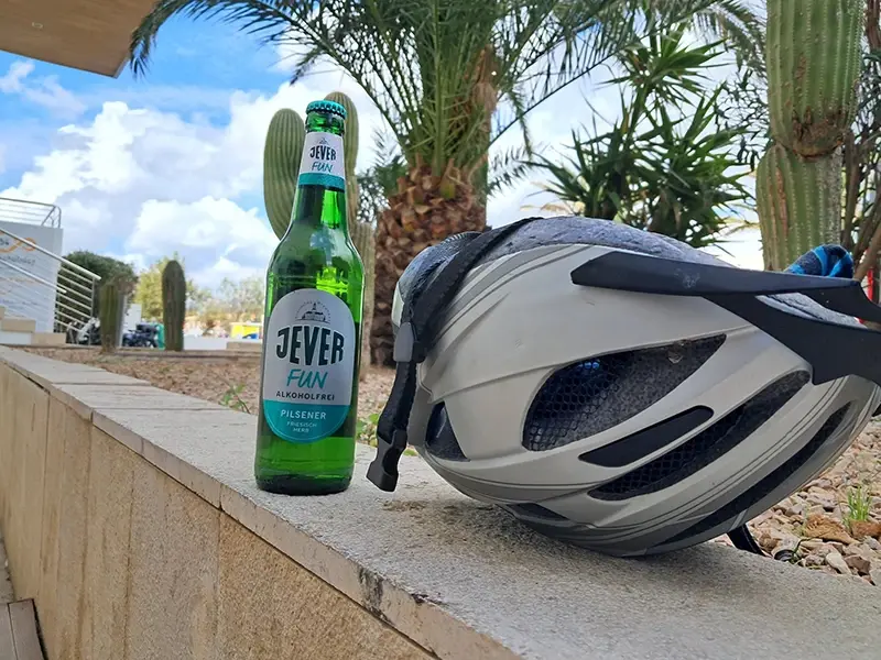 Ein Fahrradhelm liegt auf einer Mauer neben einer Flasche Jever Fun. Im Hintergrund sind Palmen zu sehen.