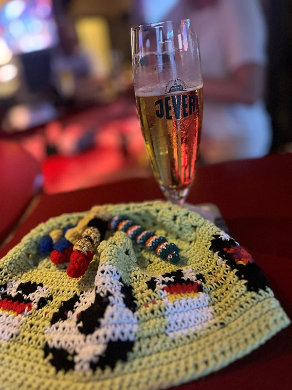 Ein kühles Glas Jever Bier auf einem Tisch, daneben eine bunte, gehäkelte Mütze mit verschiedenen Fussball-Motiven.