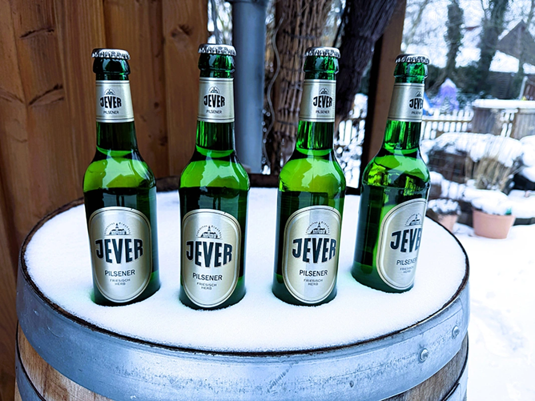Vier Flaschen Jever Pilsener stehen auf einem schneebedeckten Holzfass. Der Hintergrund zeigt eine winterliche Gartenszene.