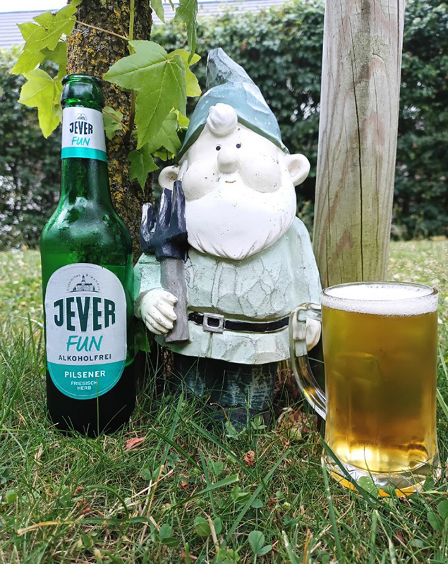 Eine Flasche Jever Fun alkoholfrei steht neben einem Gartenzwerg und einem Bierkrug auf einer Wiese. Ein Baumstamm ist sichtbar.