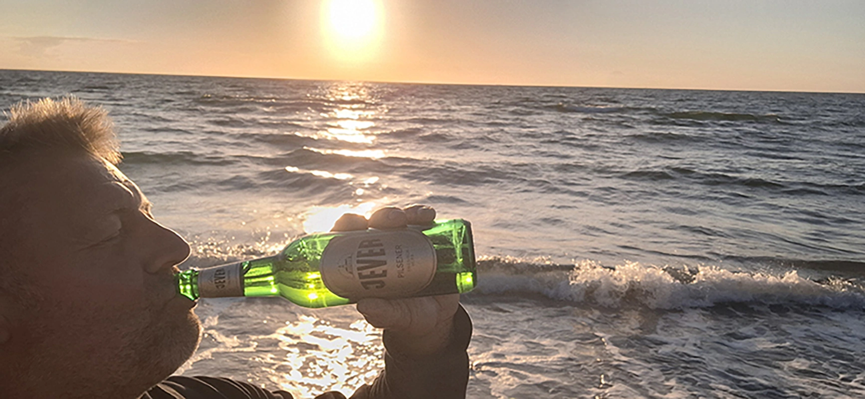 Ein Mann trinkt ein Bier am Strand, während die Sonne über dem Meer untergeht. Die Wellen brechen sanft am Ufer.