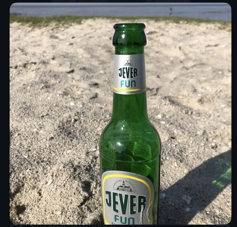 Eine Jever Fun Flasche im Vordergrund, platziert auf hellem Sand. Dahinter sieht man verschwommen Wasser und Gras.