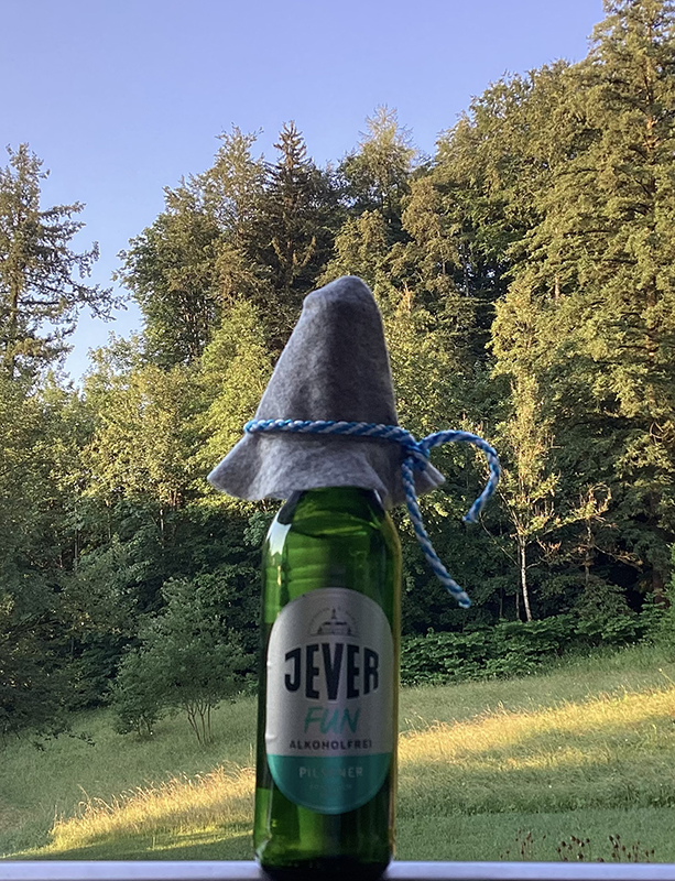 Eine Jever Fun Bierflasche mit grauem Filzhut steht auf einer Wiese, umgeben von dichten Bäumen und einem strahlend blauen Himmel.