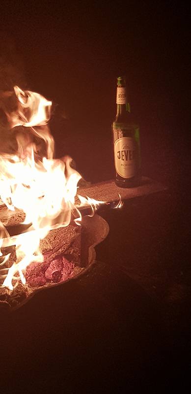 Ein Lagerfeuer brennt hell in der Dunkelheit. Daneben steht eine Flasche Jever Pilsener auf einem Stein.