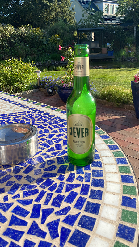 Eine Flasche Jever Pilsener steht auf einem Mosaiktisch im Garten. Im Hintergrund sind Pflanzen und ein Haus zu sehen.