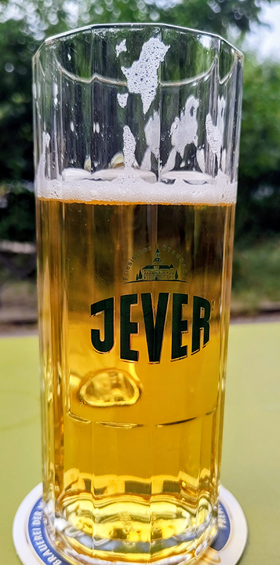 Ein halbvolles Glas mit Jever-Bier steht auf einem Bierdeckel. Im Hintergrund sind unscharfe grüne Bäume zu sehen.