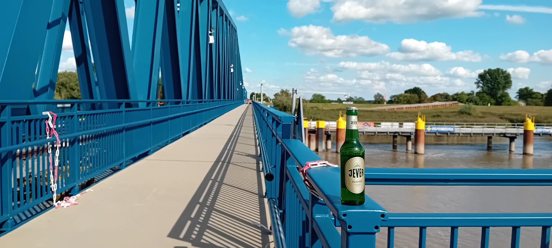 Eine blaue Brücke erstreckt sich über einen Fluss. Auf dem Geländer steht eine Jever Bierflasche. 
