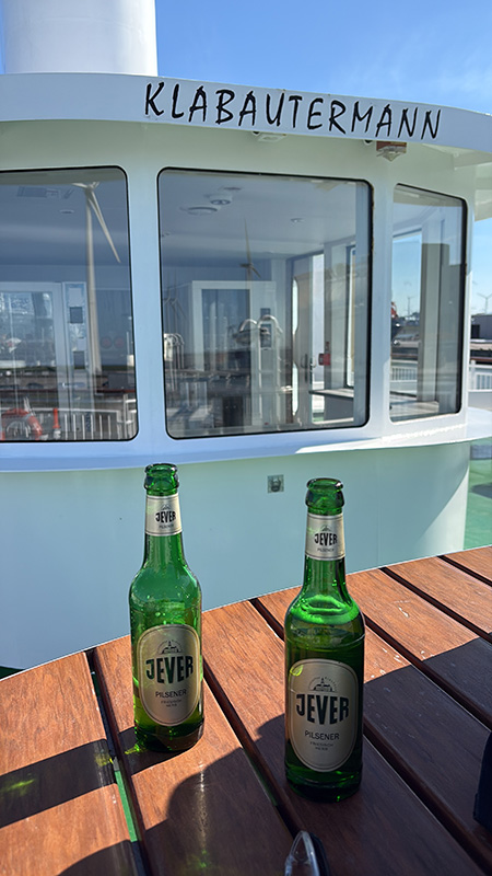 Ein Holztisch mit zwei Jever-Bierflaschen. Dahinter ein weißes Boot mit großen Fenstern und der Aufschrift 
