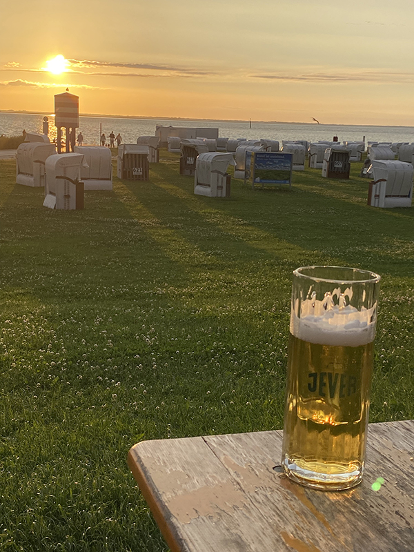 Ein Glas Jever-Bier auf einem Holztisch vor Strandkörben, während die Sonne über dem Meer untergeht.