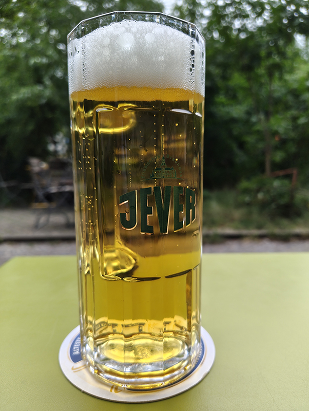 Ein großes Glas Jever-Bier mit Schaumkrone steht auf einem Bierdeckel. Im Hintergrund sind grüne Bäume zu sehen.