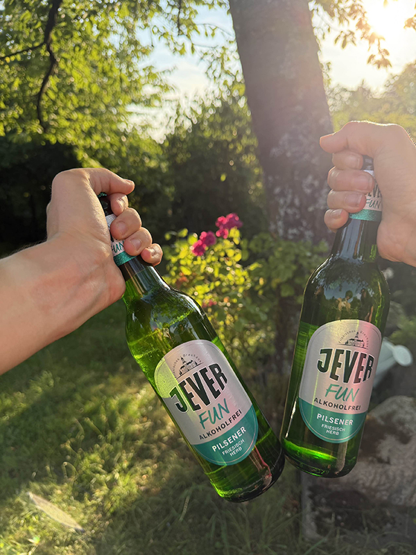 Im Garten stoßen zwei Personen mit Jever Fun Flaschen an. Die Sonne scheint, und bunte Blumen sind im Hintergrund zu sehen.