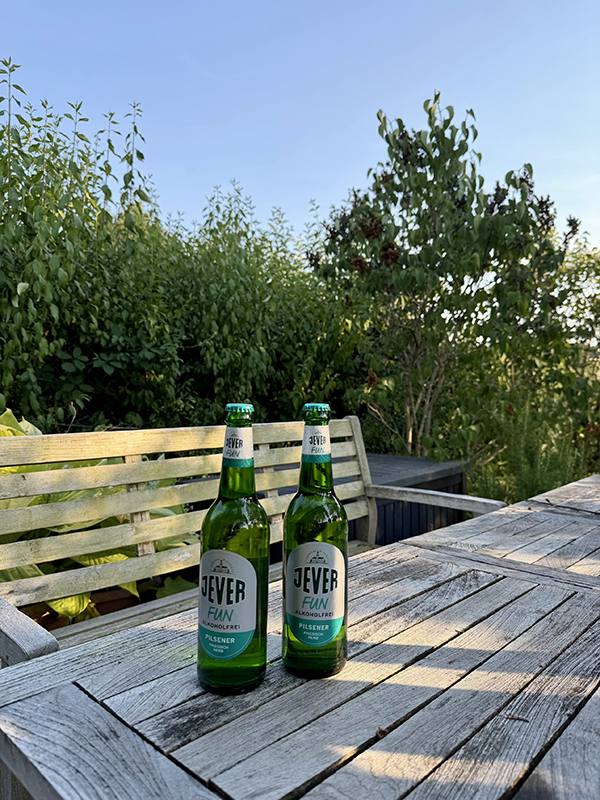 Zwei Flaschen Jever Fun Pilsener auf einem alten Holztisch. Der Garten ist von dichten Büschen und Bäumen umgeben.