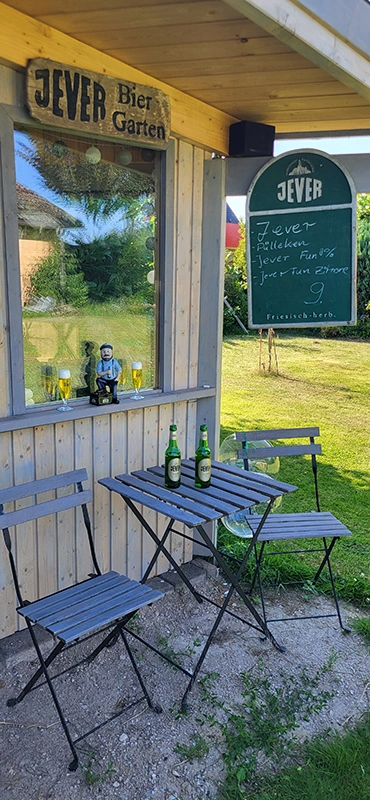 Holzhütte mit Jever-Logo und Biergarten-Schild. Zwei Bierflaschen auf dem Tisch, umgeben von grüner Wiese und Bäumen.