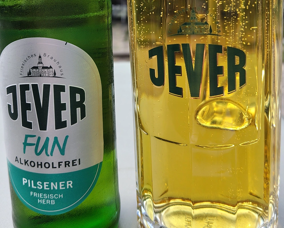 Eine grüne Flasche Jever Fun alkoholfreies Pilsener steht neben einem gefüllten Glas mit dem Jever-Logo und sprudelndem Bier.