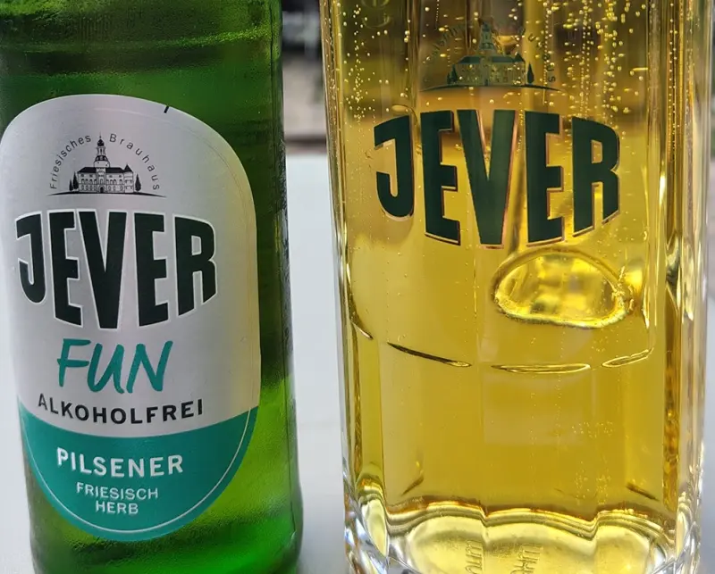 Eine grüne Flasche Jever Fun alkoholfreies Pilsener steht neben einem gefüllten Glas mit dem Jever-Logo und sprudelndem Bier.