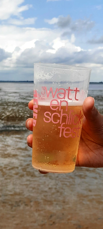 Ein Plastikbecher mit Getränk wird von einer Hand gehalten. Im Hintergrund ist ein Strand mit Wolken am Himmel zu sehen. 
