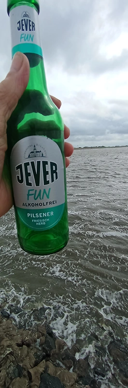 Eine Hand hält eine Flasche Jever Fun alkoholfreies Pilsener vor einer grauen, welligen Meereskulisse. 