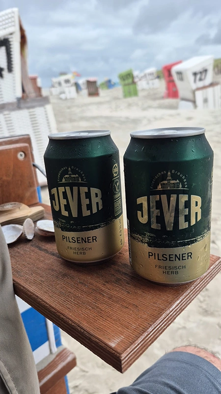 Zwei Dosen Jever Pilsener stehen auf einem Holztisch am Strand. Im Hintergrund sind Strandkörbe und bewölkter Himmel zu sehen. 