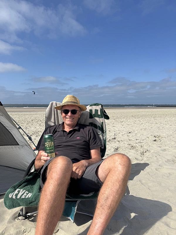 Ein Mann sitzt entspannt am Strand in einem Liegestuhl, trägt einen Strohhut und hält eine Dose Jever Bier in der Hand.