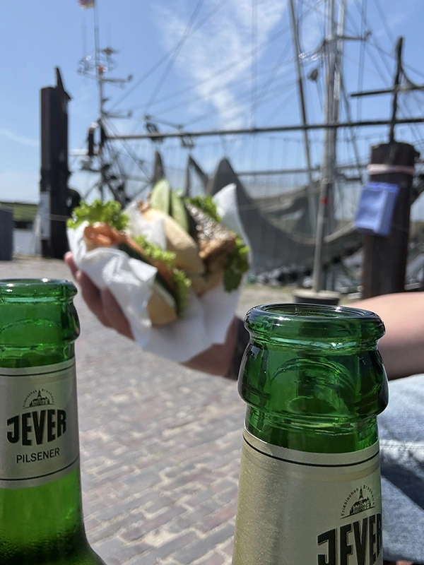 Zwei geöffnete Jever-Bierflaschen im Vordergrund, im Hintergrund eine Hand mit einem Fischbrötchen vor einem Segelschiff.