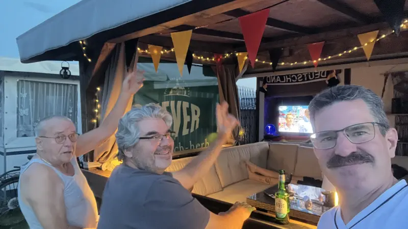 Drei Männer sitzen in einem Pavillon mit Lichterketten, schauen Fußball im TV und genießen Bier. Deutschland-Deko hängt oben.
