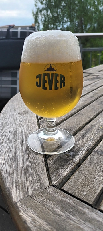 Ein Glas Jever Bier mit Schaum steht auf einem Holztisch. Der Hintergrund zeigt eine grüne Landschaft und einen bewölkten Himmel.