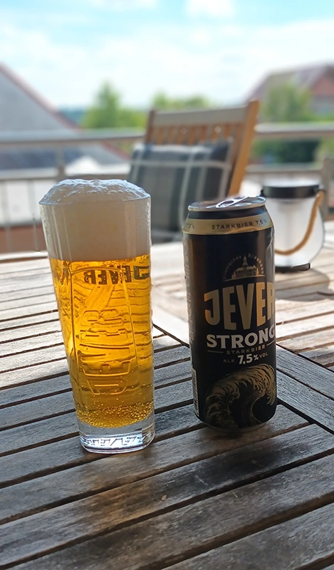 Ein Glas mit Schaumkrone und eine Dose Jever Strong Bier stehen auf einem Holztisch im Freien bei sonnigem Wetter.