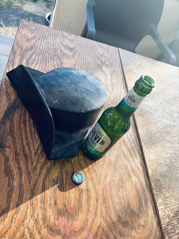 Eine grüne Bierflasche und ein schwarzer Hut liegen auf einem Holztisch. Der Kronkorken liegt daneben.