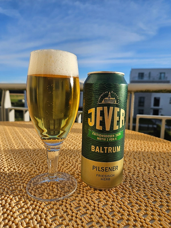 Eine grüne Jever-Bierdose und ein gefülltes Bierglas auf einem Tisch mit geflochtener Oberfläche, im Hintergrund Gebäude.